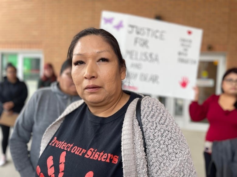 A woman in a sweater overtop of a shirt that says "Protect Our Sisters" stands in front of a person holding a sign that says "Justice for Melissa."