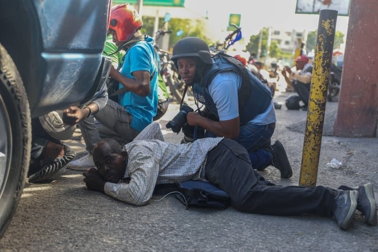 A man lays on the sidewalk while another man crouches above him with a gun as they hide behind a car avoiding gunfire.