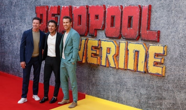 Three men stand in front of a wall that says "Deadpool & Wolverine" on it. The men are all wearing open suits or a blazer and pant combo, without ties.