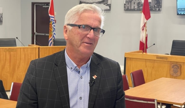 A Caucasian man with white hair, glasses, and a dark striped blazer stands in a hall with Canadian flags behind him.