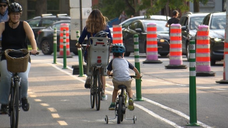 Picture of a bike lane with four bikers riding. Two bikers on the left lane are heading toward the camera. On the right lane, a boy is seen riding a bike with training wheels. He's wearing a blue helmet. He follows behind a woman biking without a helmet. A young child is sitting on a bike seat behind her, wearing a helmet.