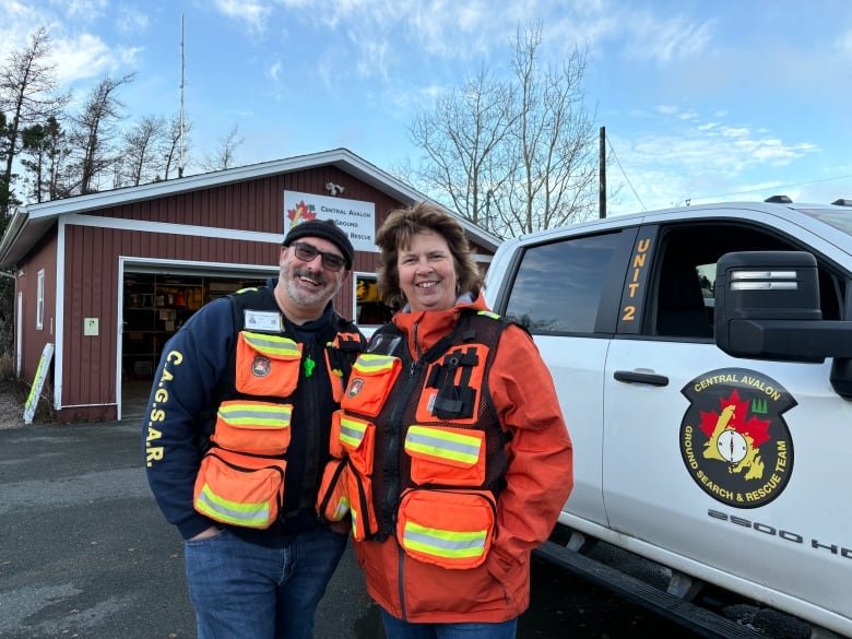 Two people wearing safety vests stand in front of a truck and a garage.