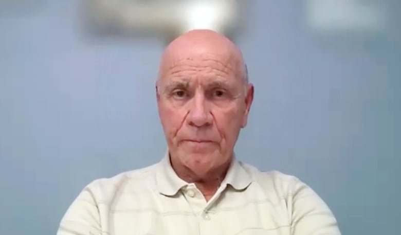 A white bald man is seen on a video call with an office wall blurred behind him