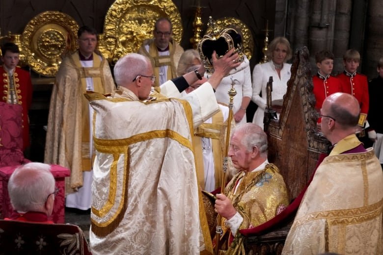 A man in religious garments stands over another man with his hands in the gesture of placing a crown atop the men's head.