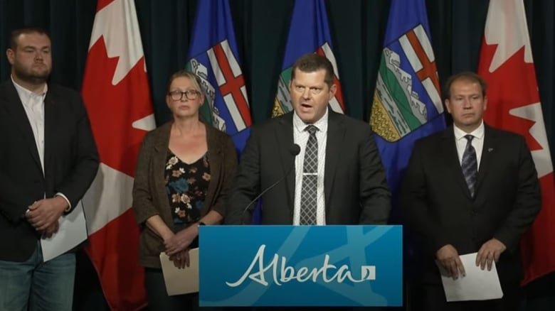 Three men and a woman stand at a blue podium that reads ALBERTA in cursive font.