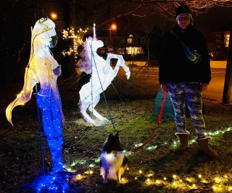 Six-feet-tall plastic decorations that are lit with bright internal lands and anchored with rods. Nearby, a woman in winter clothes stands with a dog on a leash.