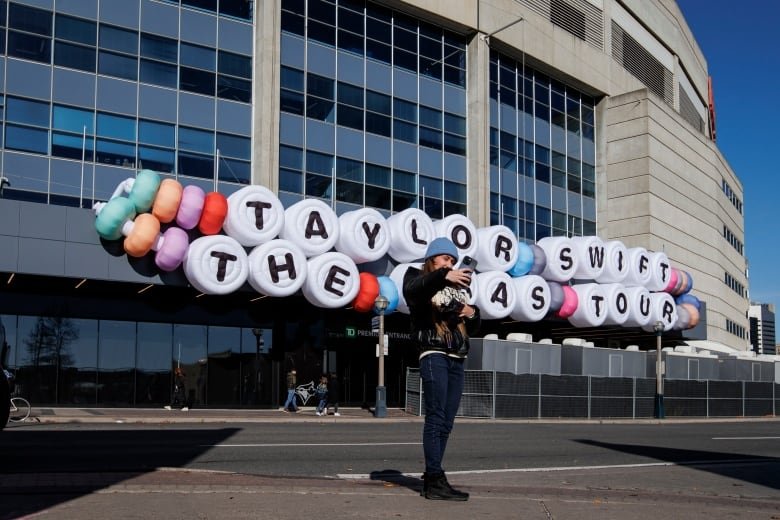 A woman takes a selfie in front of Rogers Centre stadium where hangs a giant friendship bracelet sign that reads: Taylor Swift The Eras Tour.