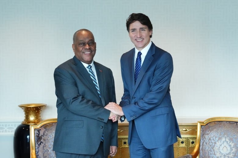 Prime Minister Justin Trudeau and Haiti Prime Minister Garry Conille shake hands. Both men are wearing suits and are shown standing in front of two arm chairs.