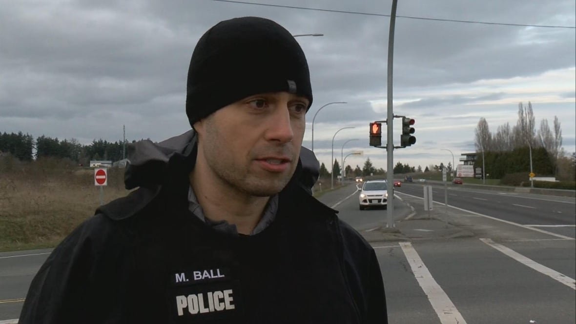 A police officer wearing a tuque and a name badge reading 'M. Ball' speaks at a traffic intersection.