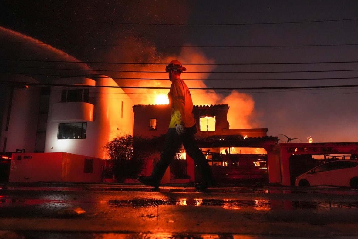 a firefighter walks in front of a burning building at night.