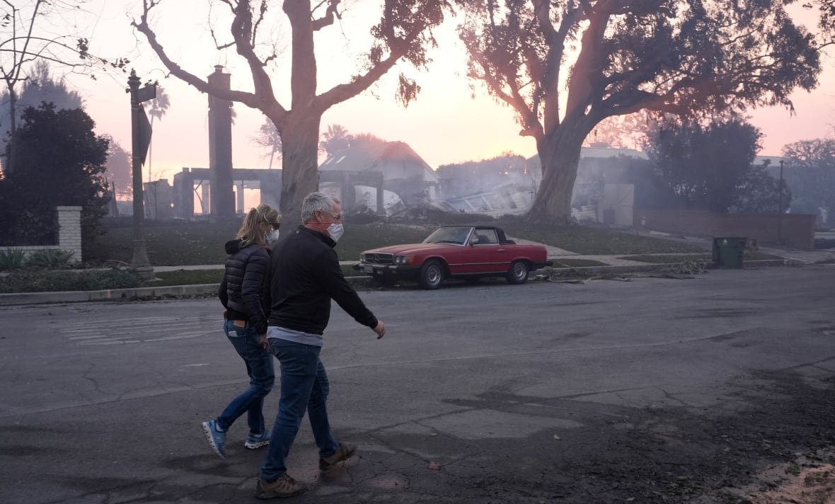 two people walk on a road, both wearing medical masks over their noses and mouths. there is a car parked on the street in front of burned buildings