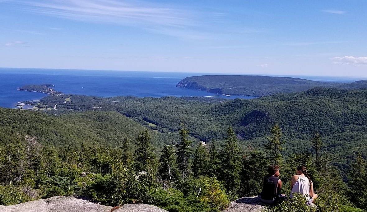 Two people sit on a rock overlooking a lush green valley and the ocean in Cape Breton Highlands National Park.