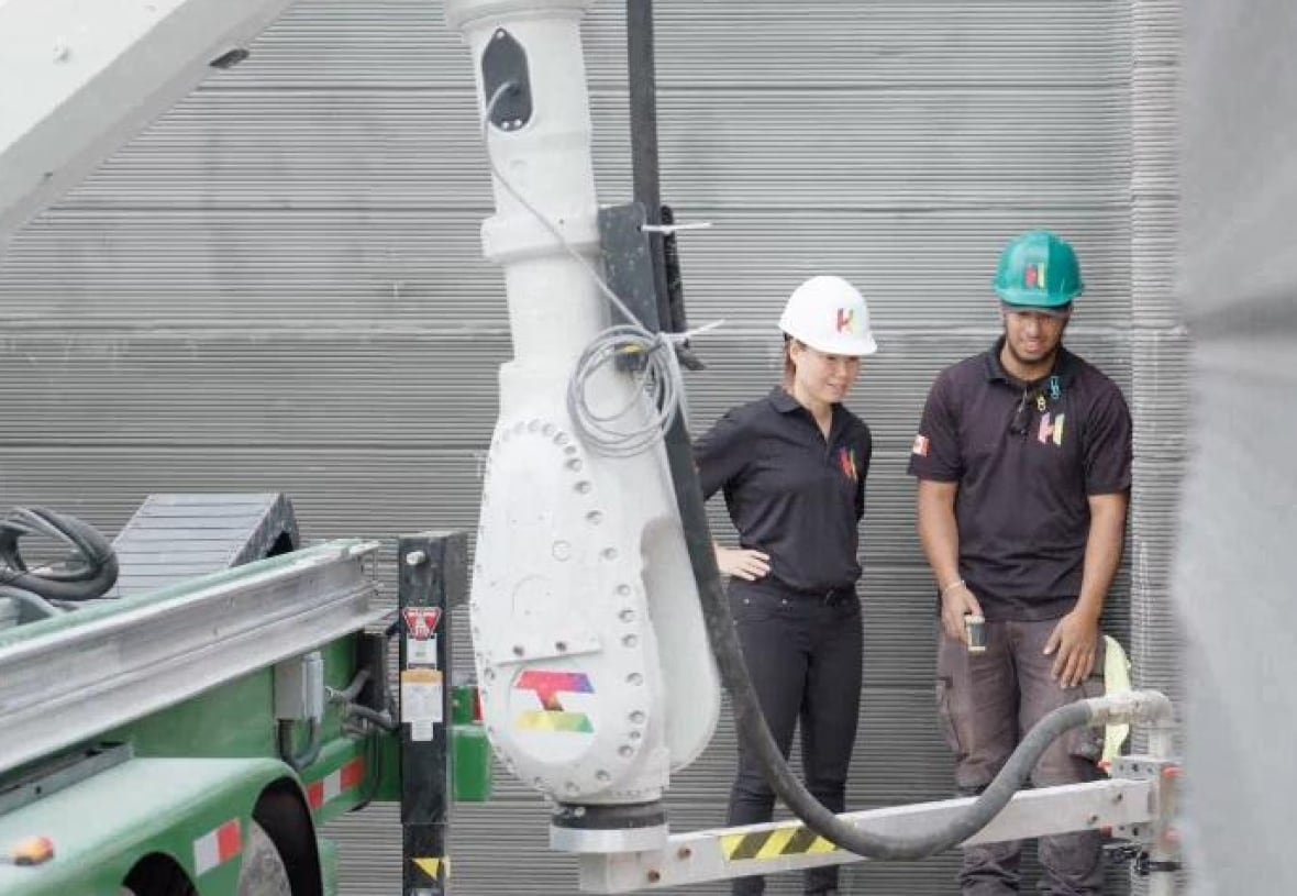 A woman in a white hard hat and man in a blue hard hat stand near a concrete wall beong poured by a large robotic arm equiped with a tube and hose.