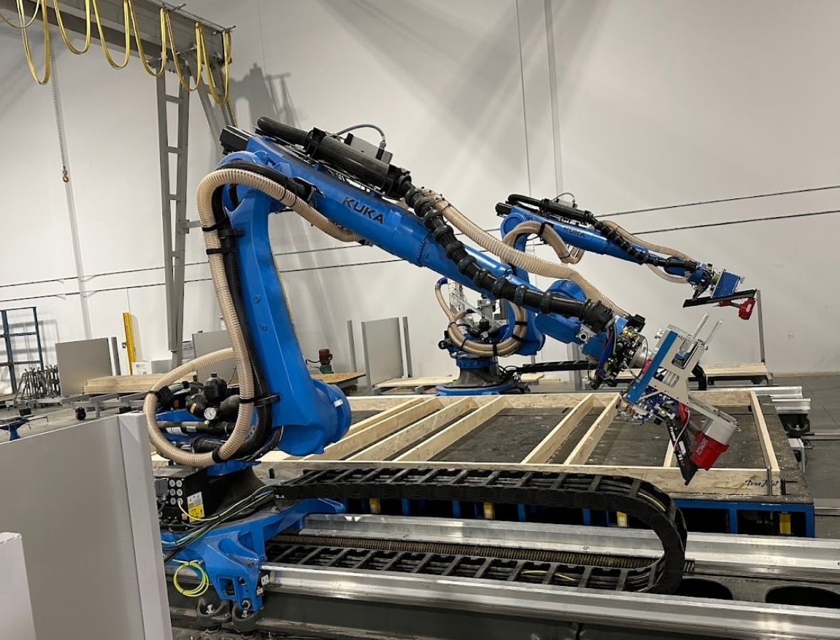 Two big blue robotic arms loom over the wood frame wall of a house which lays on a large table. The arms hold red nail guns to nail peices together.