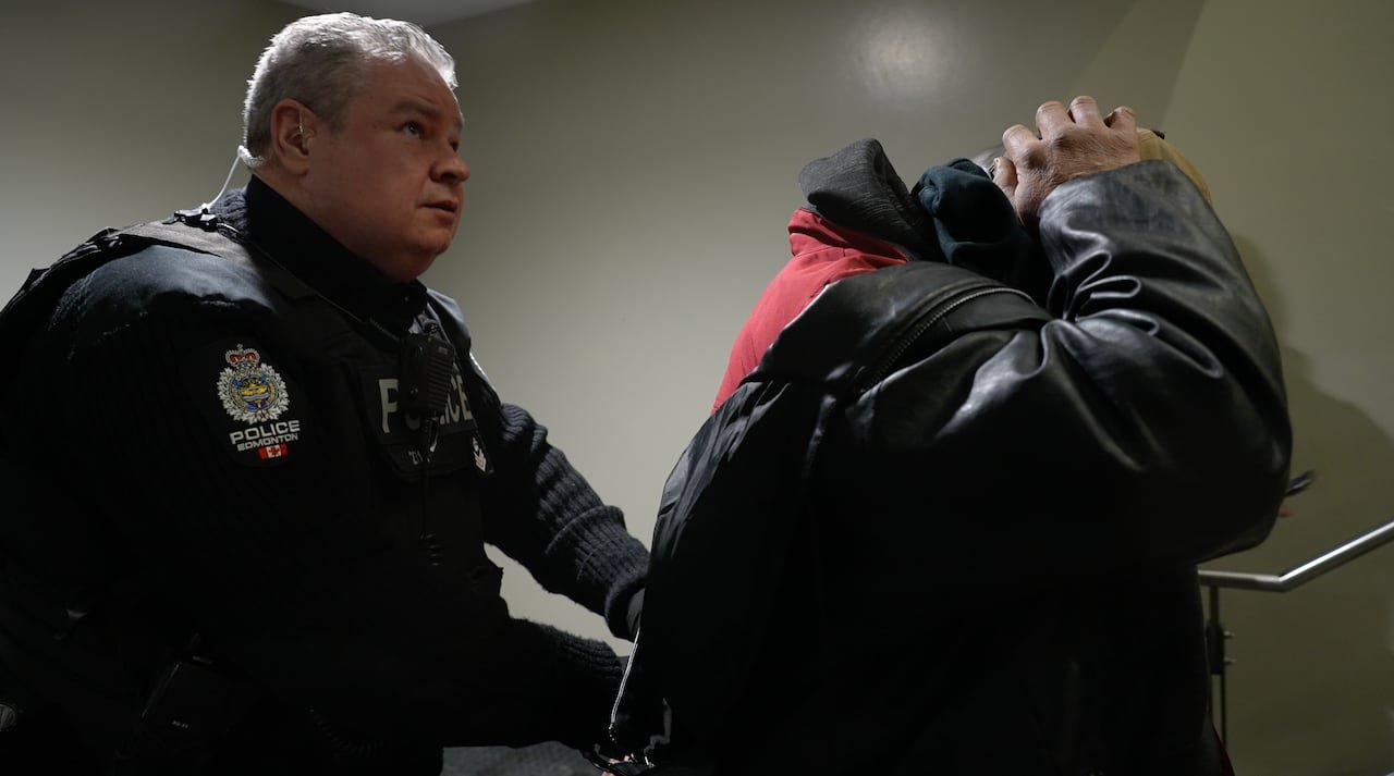 A police officer stands behind a man who has his hands on his head.
