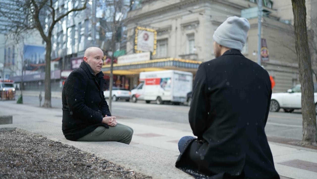 Two people sit on a ledge and speak.