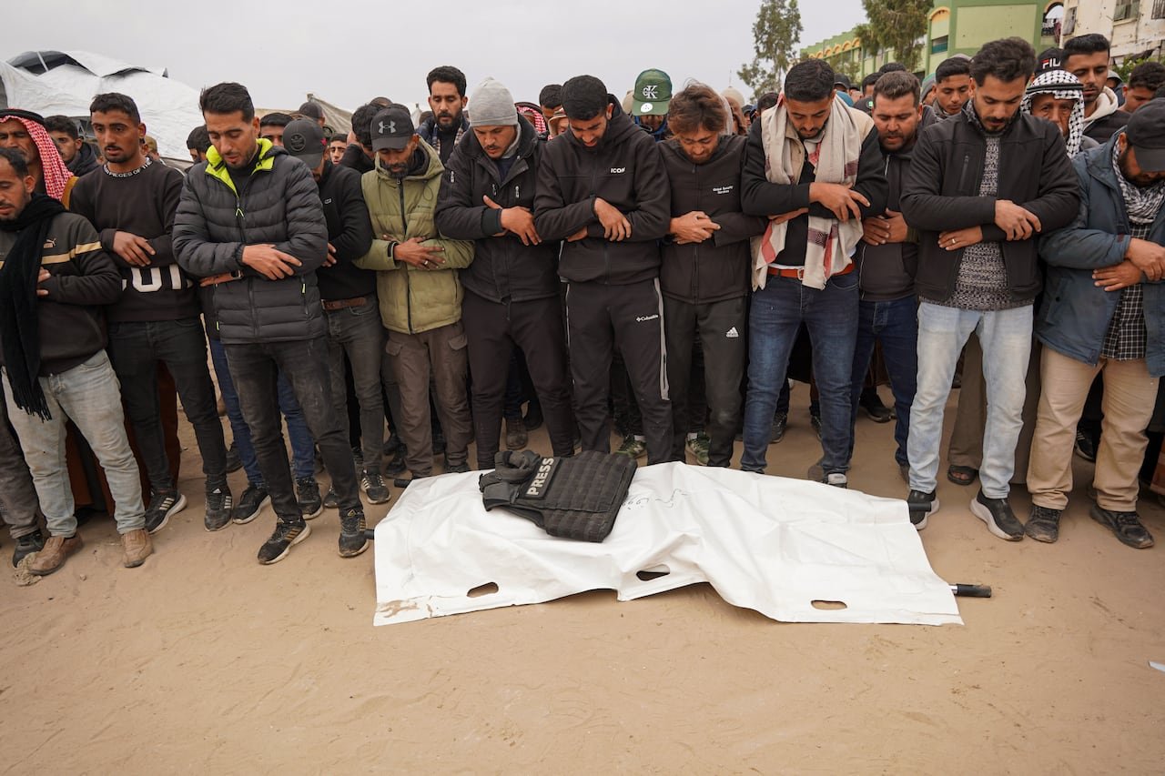 default-472 A group of men standing near a shrouded dead body adorned with a bulletproof vest labeled 'Press.'